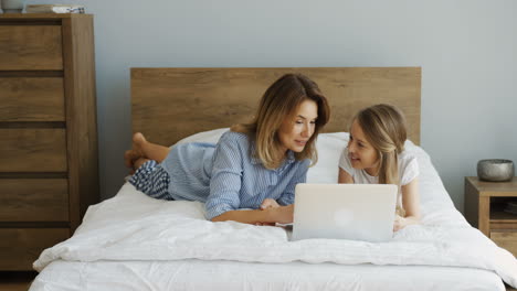 Cheerful-smiled-mother-and-litle-daughter-resting-after-waking-up-in-the-morning-on-the-unmade-bed-in-front-of-the-laptop-screen.-Indoors